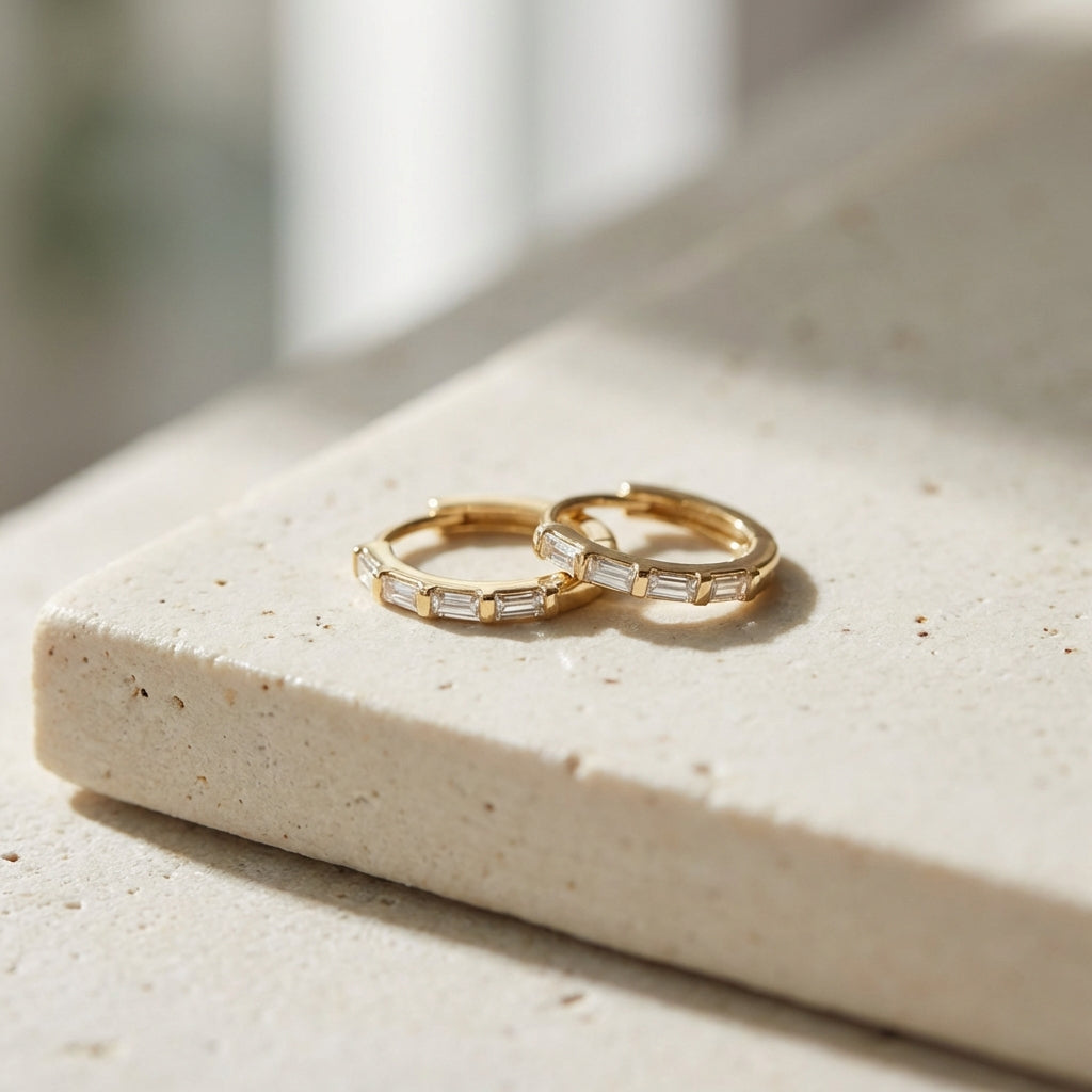 Artistic studio scene with 14K gold baguette diamond huggie earrings displayed on a textured surface, emphasizing their delicate craftsmanship.