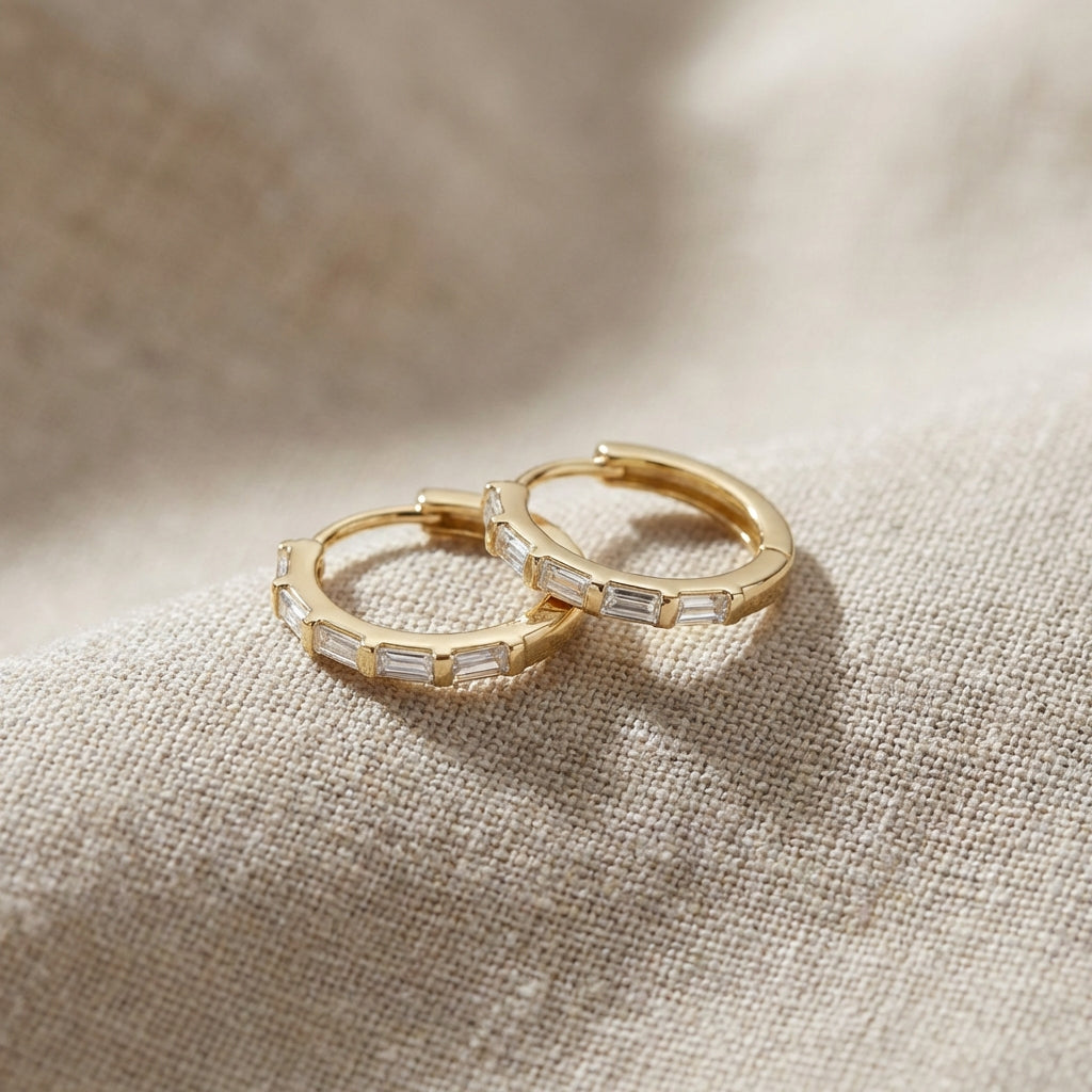Close-up of delicate 14K gold baguette diamond huggie earrings highlighting their fine details and polished surface against a textured fabric backdrop.